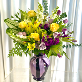 Yellow and purple mixed bouquet in a dark vase with a lavender ribbon