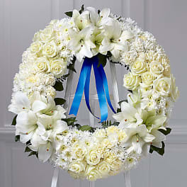 White floral wreath with roses and lilies on a stand, tied with a blue ribbon