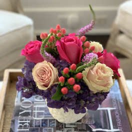 Pink and cream roses arranged in a small white vase with purple flowers.