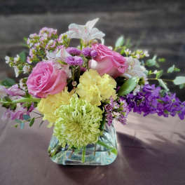Small mixed arrangement of pink roses, yellow carnations and purple blooms in a clear glass cube vase