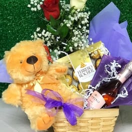 Gift basket with teddy bear, red and white roses, chocolates, and a pink sparkling drink wrapped with purple ribbon