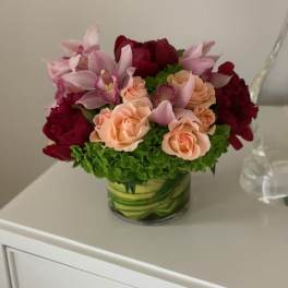 Compact bouquet of pink orchids, peach roses, and deep red blooms in a glass vase