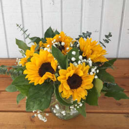 Arrangement of bright yellow sunflowers and white filler flowers in a clear glass vase on a wooden table