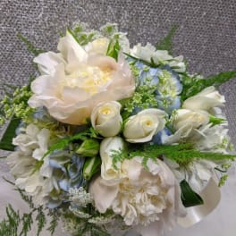 Round bouquet of ivory peonies, white roses, and pale blue flowers in a glass vase.