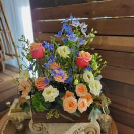 Mixed bouquet of peach, cream, and pink roses with blue filler flowers in a wooden box