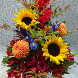 Tall mixed arrangement with sunflowers, red roses, orange dahlias, and purple iris in a clear glass vase