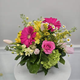 Bright mixed arrangement with hot pink gerbera daisies, pink rose, tulips, and yellow blooms in a clear glass vase