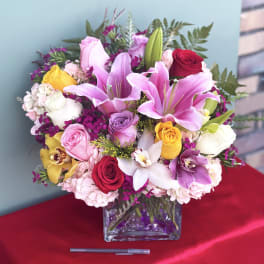 Mixed bouquet of pink lilies, roses, and orchids in a square glass vase