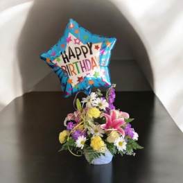 Birthday balloon above a small mixed flower arrangement in a basket