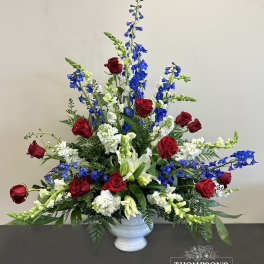 Tall red, white, and blue arrangement with roses, lilies, and delphinium in a white urn vase