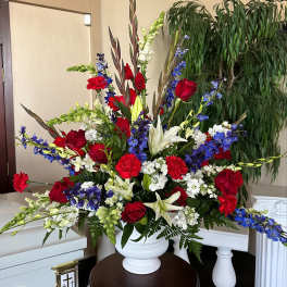 Tall red, white, and blue funeral arrangement with roses, lilies, carnations, and delphinium in a white urn.