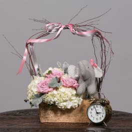 Pink roses and white hydrangeas in a wooden basket with a knitted elephant and clock