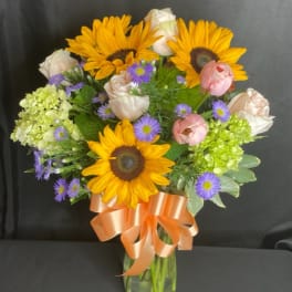 Bouquet of sunflowers, roses, and purple daisies in a glass vase with a peach ribbon
