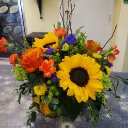 Bright mixed bouquet with sunflowers, orange roses, and purple accents in a low vase