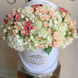 Large hatbox arrangement of pastel roses and white hydrangeas in a white container.