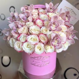 Round pink hat box filled with cream roses edged in pink and pale pink orchids, with a small card tucked in.