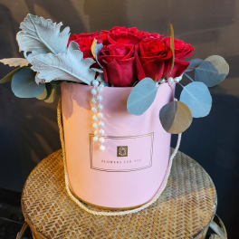 Red roses arranged in a pink hatbox with pearl strands and blue-gray foliage.