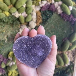 Hand holding a purple heart-shaped crystal against a floral wall