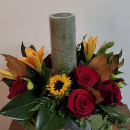 Red roses and orange lilies arranged around a tall candle in a glass bowl
