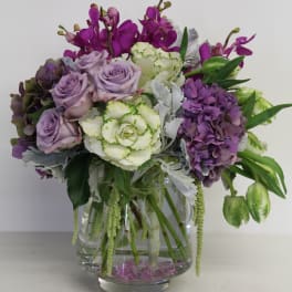 Lavender roses, purple hydrangea, orchids, and green-white blooms in a clear glass vase