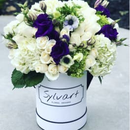 White and purple floral arrangement in a round hat box