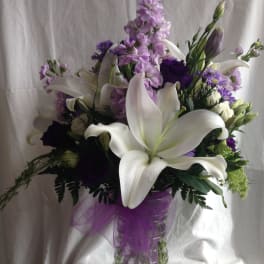 White lilies and purple flowers arranged in a glass vase with a purple ribbon.