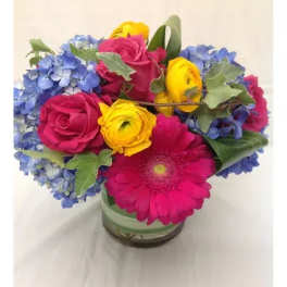 Bright bouquet of pink roses and gerbera daisy, yellow ranunculus, and blue hydrangeas in a glass vase