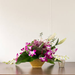 Purple orchids and lilies arranged in a gold vase with a small candle holder beside it