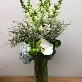 Tall white and green floral arrangement in a glass vase with blue hydrangea and orchid