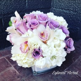 Lavender roses and white orchids arranged in a square glass vase