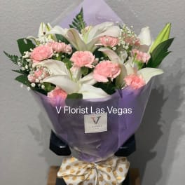 Hand-tied bouquet of white lilies and pink carnations wrapped in purple paper with a polka dot bow.