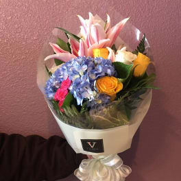 Hand-tied bouquet of pink lilies, blue hydrangea, and yellow and white roses in cream wrap with a bow.