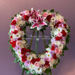 Heart-shaped standing wreath of pink lilies, roses, red carnations, and white daisies on an easel.