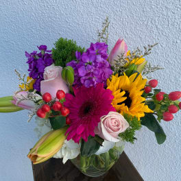 Colorful mixed bouquet with sunflowers, roses, lilies, and a gerbera daisy in a glass vase
