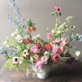 Asymmetrical garden-style arrangement of pink, coral, and white flowers with blue accents in a white vase