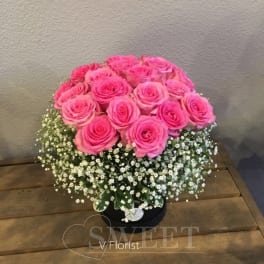 Round arrangement of bright pink roses with a collar of white filler flowers in a black hatbox.