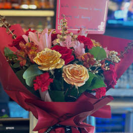 Red and gold rose bouquet with pink lilies and mixed blooms wrapped in deep red paper with a message card.