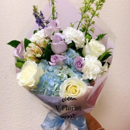 Hand-tied bouquet of white and lavender roses, blue hydrangeas, and white carnations wrapped in pastel paper.