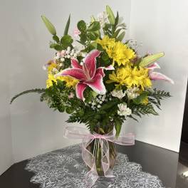 Pink lilies and yellow daisies in a glass vase with a pink ribbon