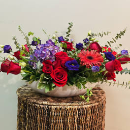 Mixed bouquet of red roses, purple blooms, and a pink gerbera daisy in a white bowl
