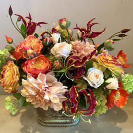 Lush centerpiece of orange, peach, and yellow blooms with burgundy accents in a decorative ceramic container