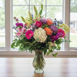 Mixed bouquet of pink lilies, roses, gerbera daisies, and white hydrangea in a clear glass vase