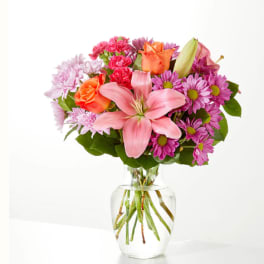 Pink lilies, orange roses, and purple daisies in a clear glass vase