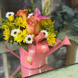 Pink watering can holding yellow and white flowers with a rose and ribbon