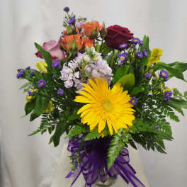 Mixed bouquet with a yellow gerbera daisy, roses, and purple filler flowers in a glass vase