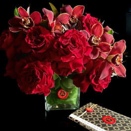 Red roses and orchids arranged in a clear square vase beside a patterned gift box