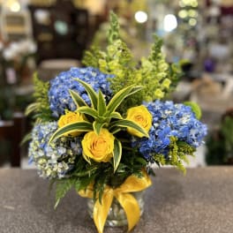 Blue hydrangeas and yellow roses in a glass vase with a yellow ribbon