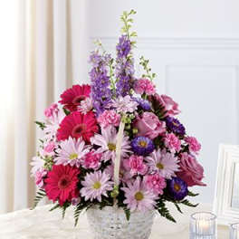 Pink and purple mixed flower basket arrangement in a white woven basket on a table