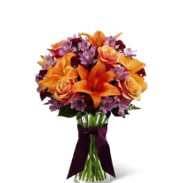 Vase arrangement of orange lilies, peach roses, and purple flowers in a clear glass vase with dark ribbon