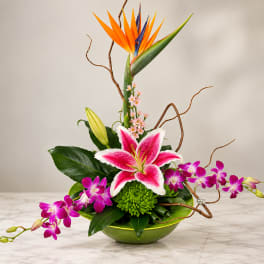 Tropical flower arrangement with pink lilies and purple orchids in a green bowl